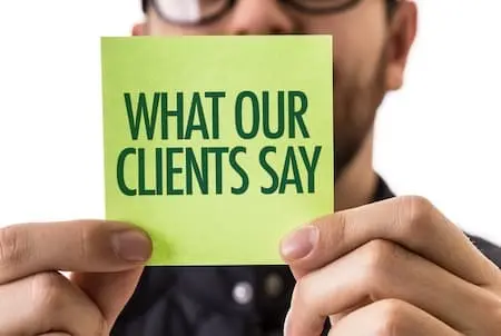 Close-up of a person holding a sticky note that reads “What Our Clients Say,” representing the importance of client testimonials for life coaches.