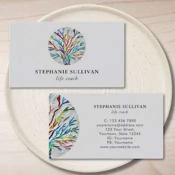 Business card example from Zazzle for a life coach featuring front and back designs with a colourful abstract tree logo, professional name, contact details, and social media links on a clean white background.