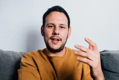 Man sitting on a couch, speaking with expressive hand gestures, illustrating a client recording a video testimonial for a life coach.